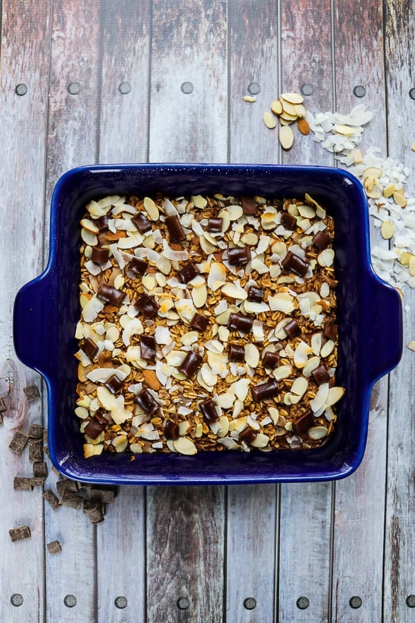 Baked chocolate oatmeal in a square baking dish topped with chocolate chunks, almonds and coconut.