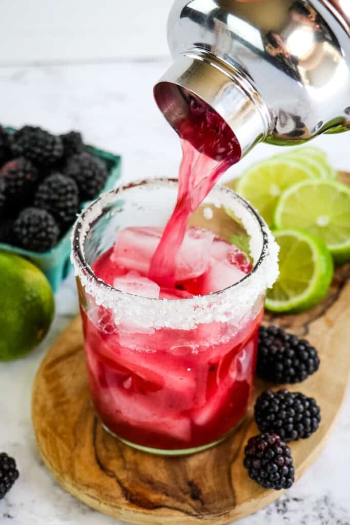Blackberry margarita being poured into a glass with a salted rim.