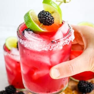 Blackberry margarita held in hand and topped with fresh blackberries and lime slice.