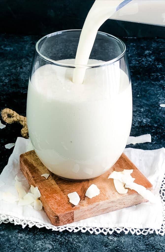 Coconut smoothie being poured into a glass with coconut pieces on the side.