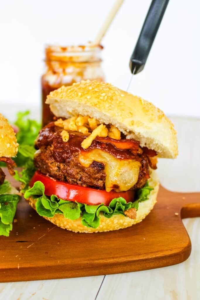 BBQ bacon burger with lettuce, tomato, cheese, sauce, bacon and onions, with knife going through the top of the burger.