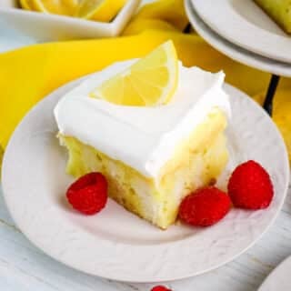 Slice of lemon poke cake topped with a lemon slice and with fresh raspberries on the side.