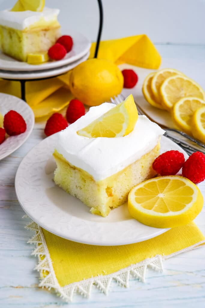 Slice of lemon poke cake on a plate with lemon slice and fresh raspberries.