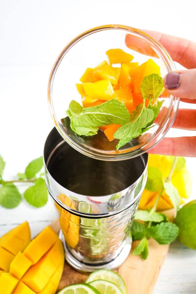 Mango pieces and mint leaved being placed into a cocktail shaker for making a mango mojito.