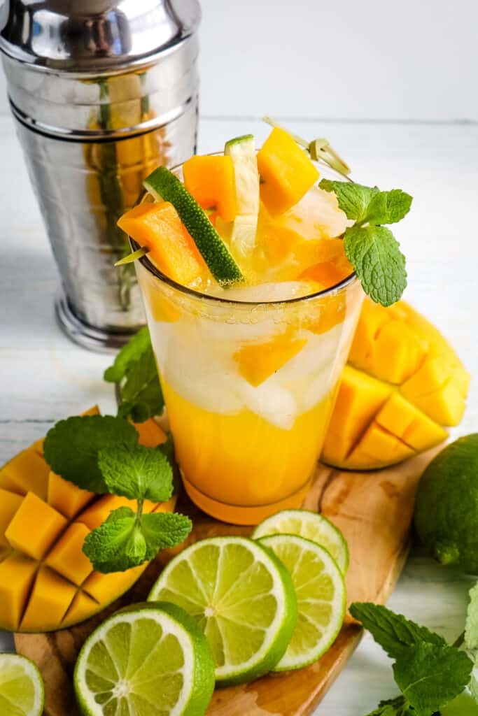 Mango mojito recipe in glass with cocktail shaker in the background and garnished with lime slices, mint and mangos on the side.