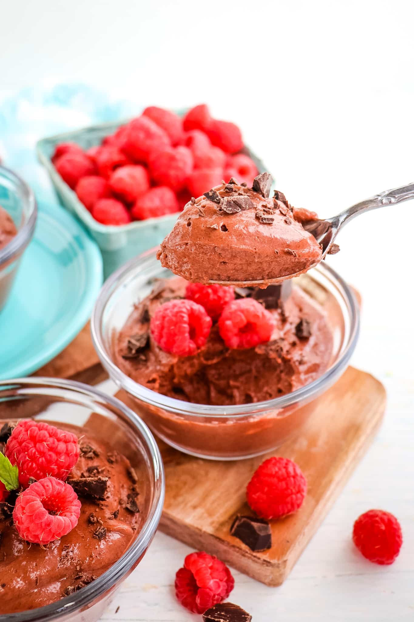 Spoon taking bite out of chocolate protein pudding, topped with chocolate chunks and fresh raspberries.