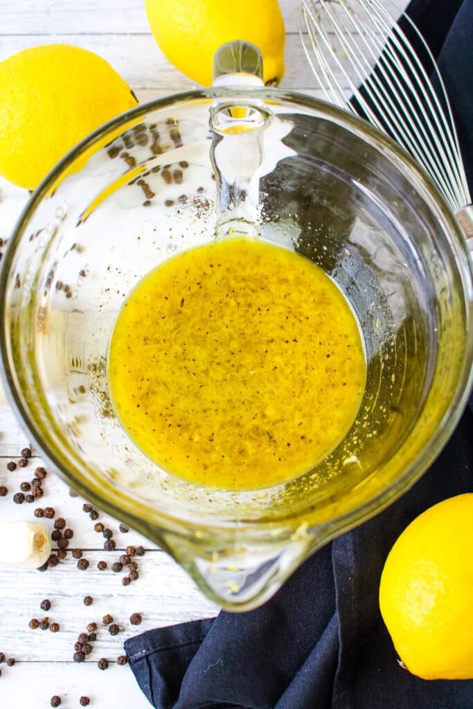 Lemon olive oil dressing in a mixing bowl with lemons and whisk on the side.