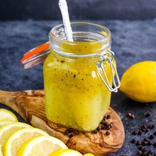 Lemon olive oil dressing in a small jar with a spoon in the jar and sliced lemons on the side.