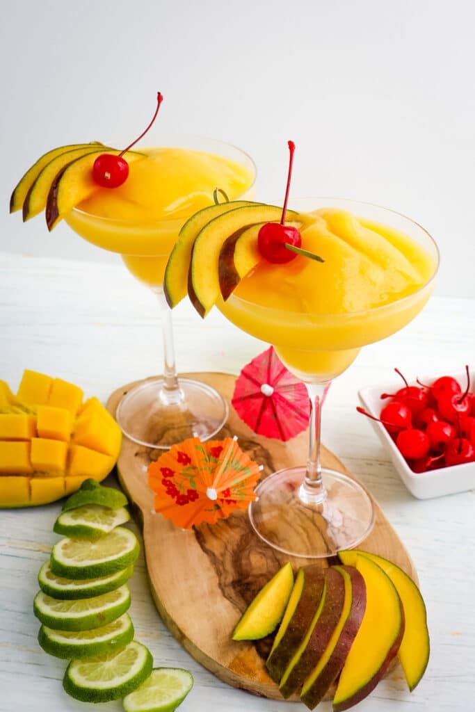 Two mango daiquiri's with lime and mango slices and maraschino cherries on the side.