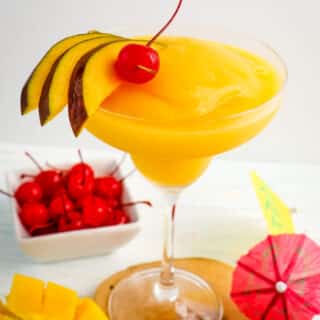 Frozen mango daiquiri in a glass topped with sliced mangoes and a maraschino cherry.