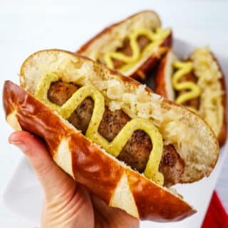 Air fryer brat in pretzel roll held in hand and topped with brown mustard and sauerkraut.