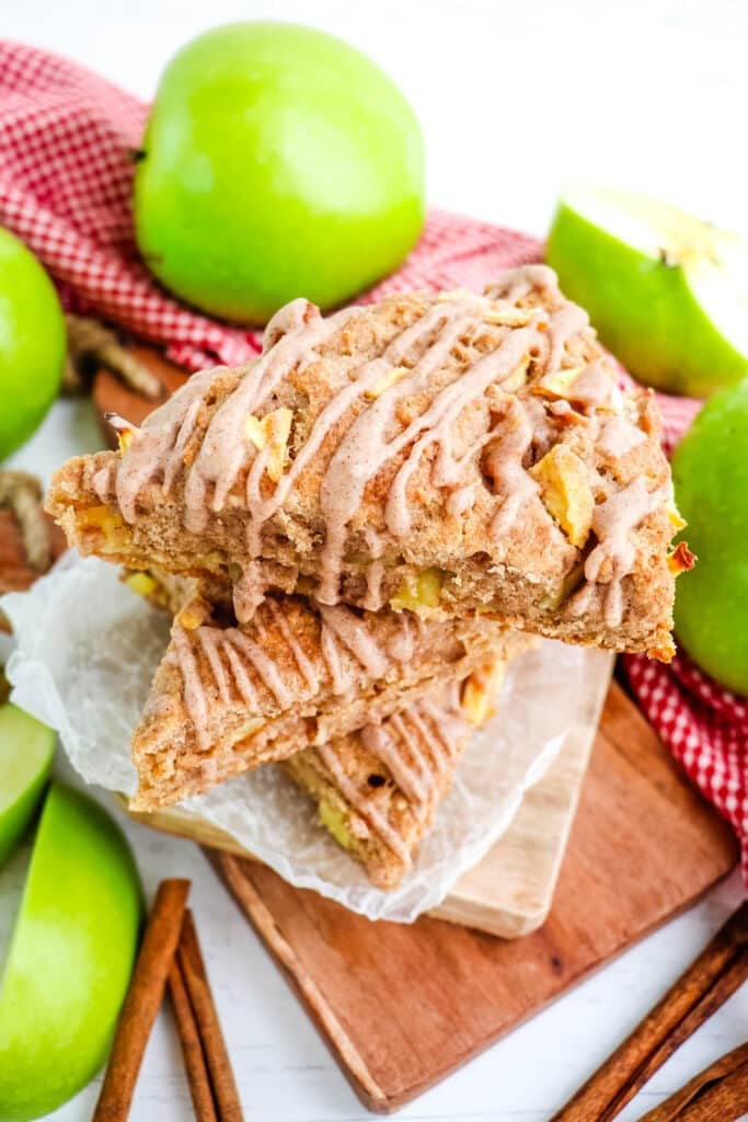 Stack of three apple scones sitting on a wooden board with green apples and cinnamon sticks in the background.