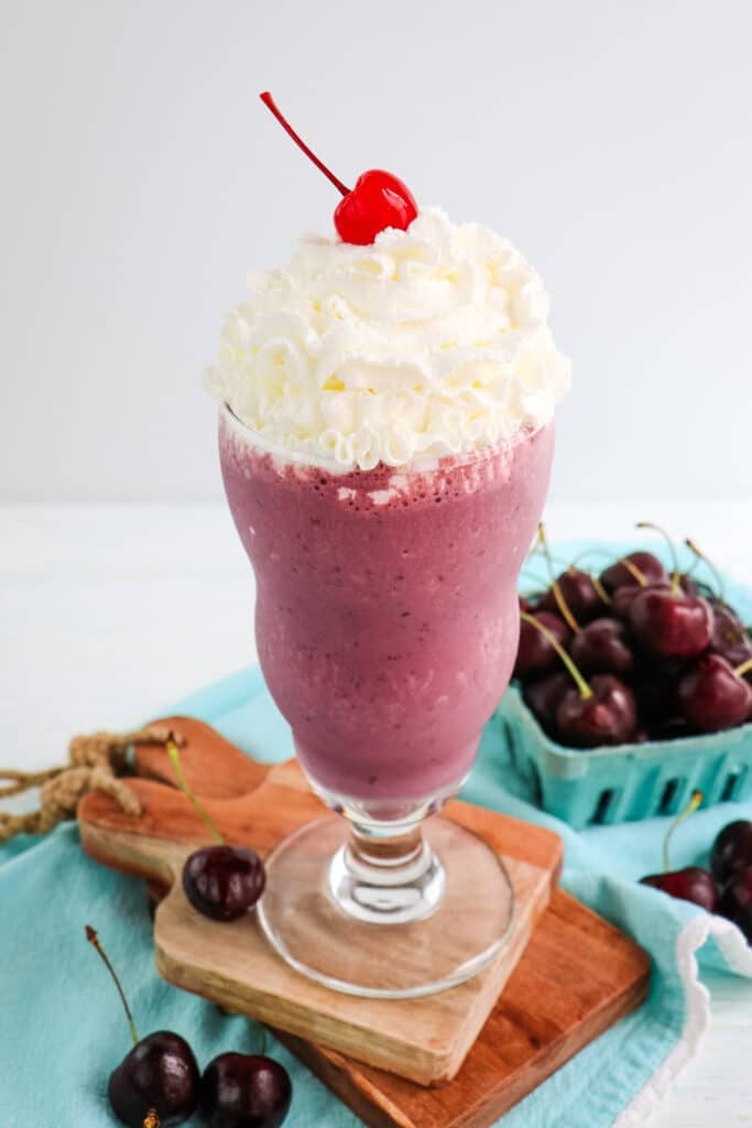 Homemade cherry milkshake in a tall glass topped with whipped cream and a maraschino cherry.