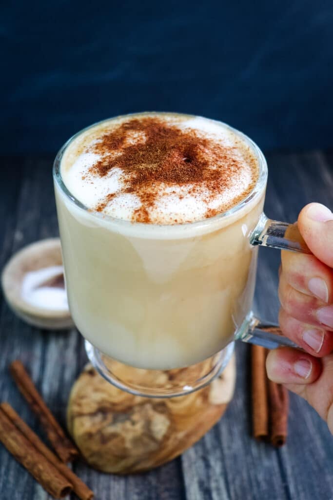 A cinnamon dolce latte held in hand in a clear mug topped with ground cinnamon.