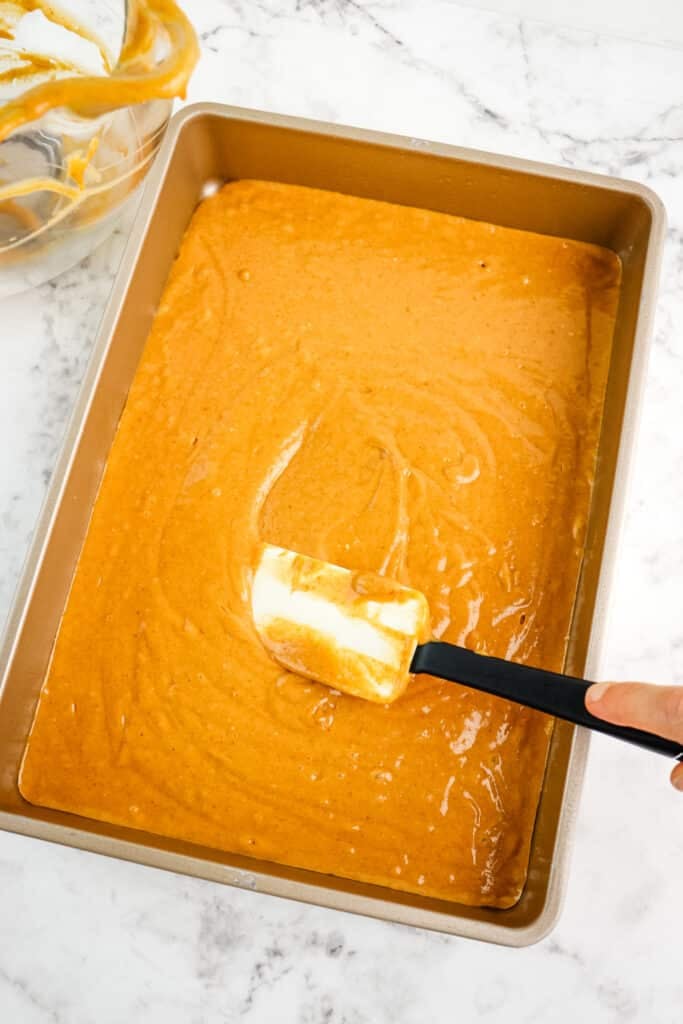 Pumpkin poke cake batter poured into a pan and batter being smoothed with a rubber scraper.