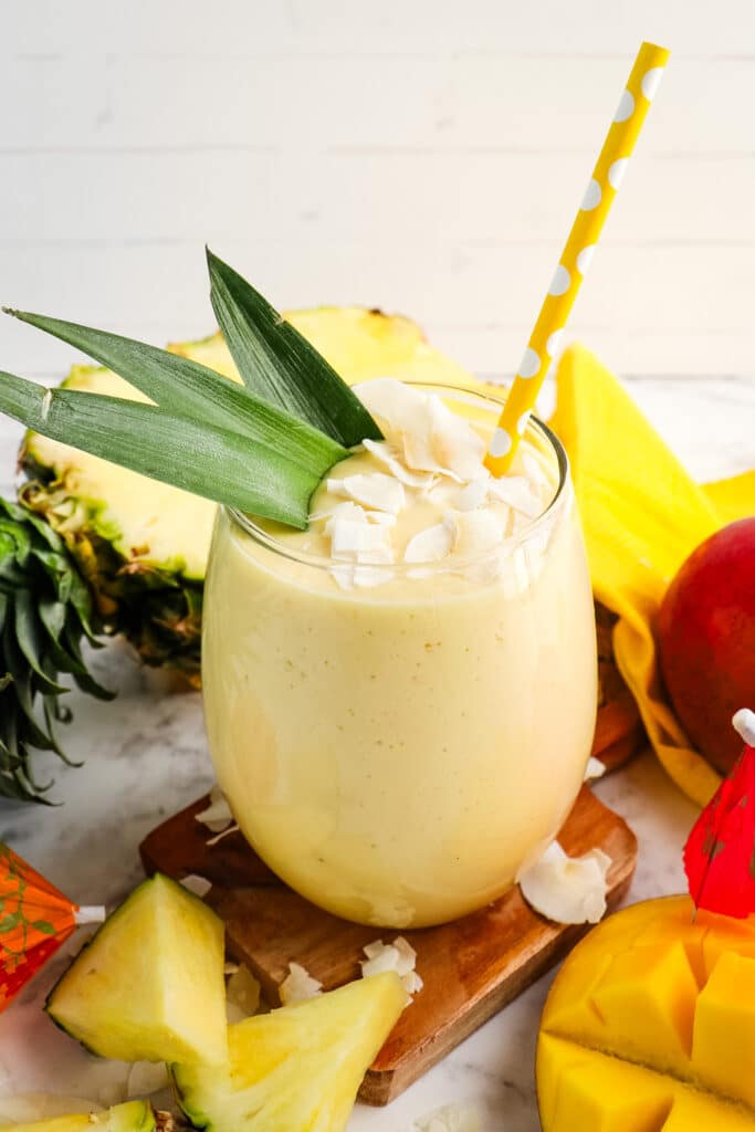 Mango pineapple smoothie garnished with pineapple leaves, coconut flakes and topped with a yellow straw.