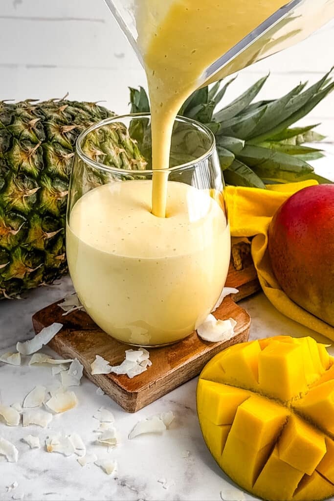 Mango smoothie being poured into a clear glass, with mango and pineapple on the side.