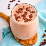 Mocha smoothie in a clear glass topped with chocolate chunks and coffee beans on the side.