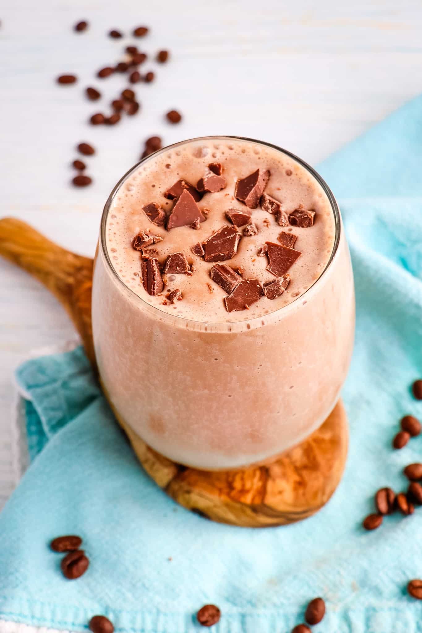 Mocha smoothie in a clear glass topped with chocolate chunks and coffee beans on the side.