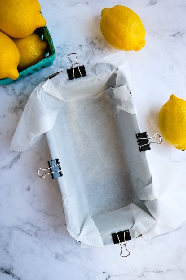 Loaf pan lined with parchment paper secured with binder clips on the sides, with lemons on the side.
