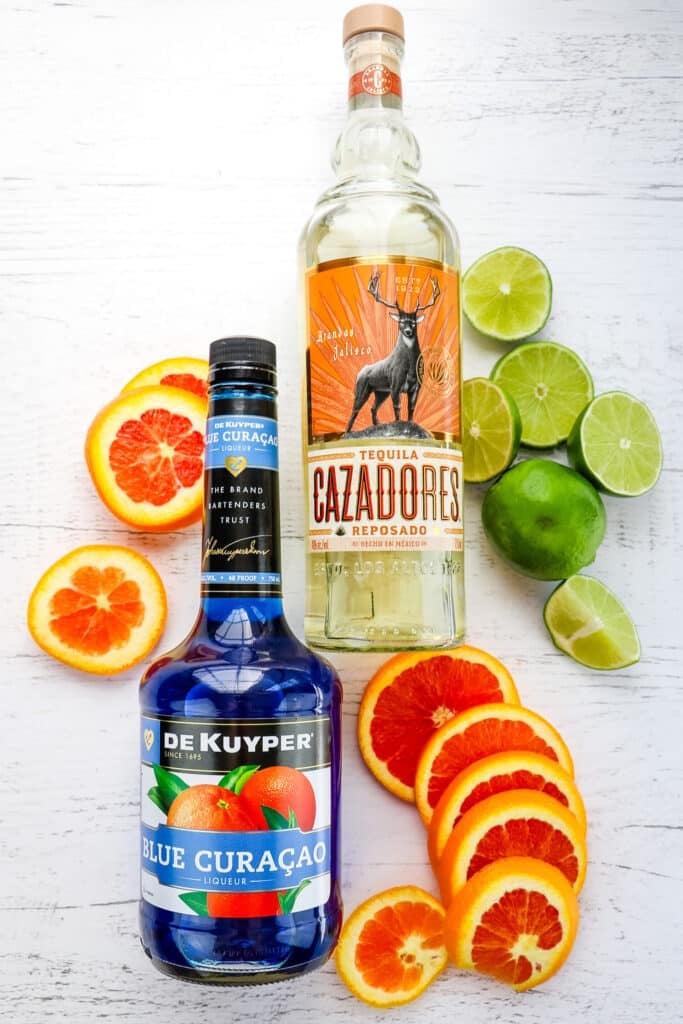 Bottles of tequila, blue curacao, lime slices and orange slices to make blue margaritas.