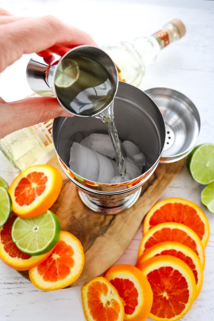 Tequila being poured into a cocktail shaker with orange slices on the side.
