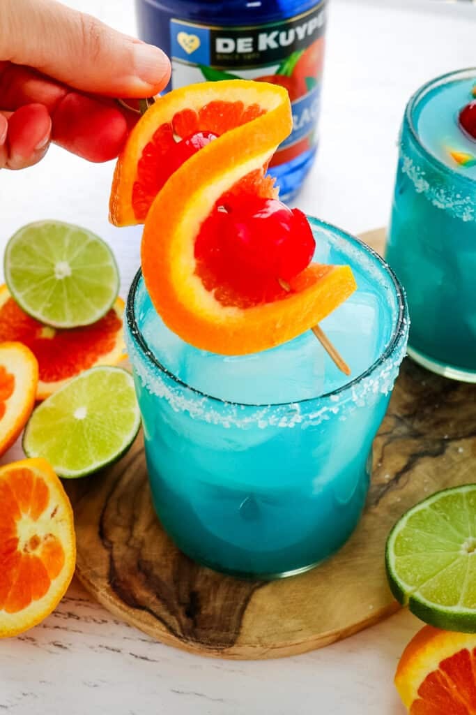 A blue margarita being topped with an orange and cherry twist garnish.