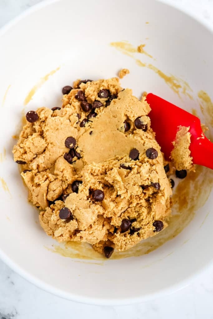 Chocolate chip cookie dough protein balls batter in a white mixing bowl with red rubber scraper on the side.