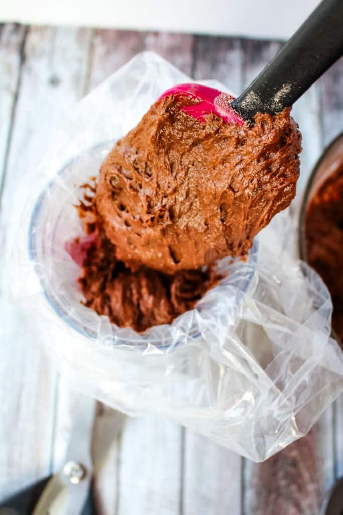 Chocolate cake batter being added to a plastic bag with a rubber scraper.