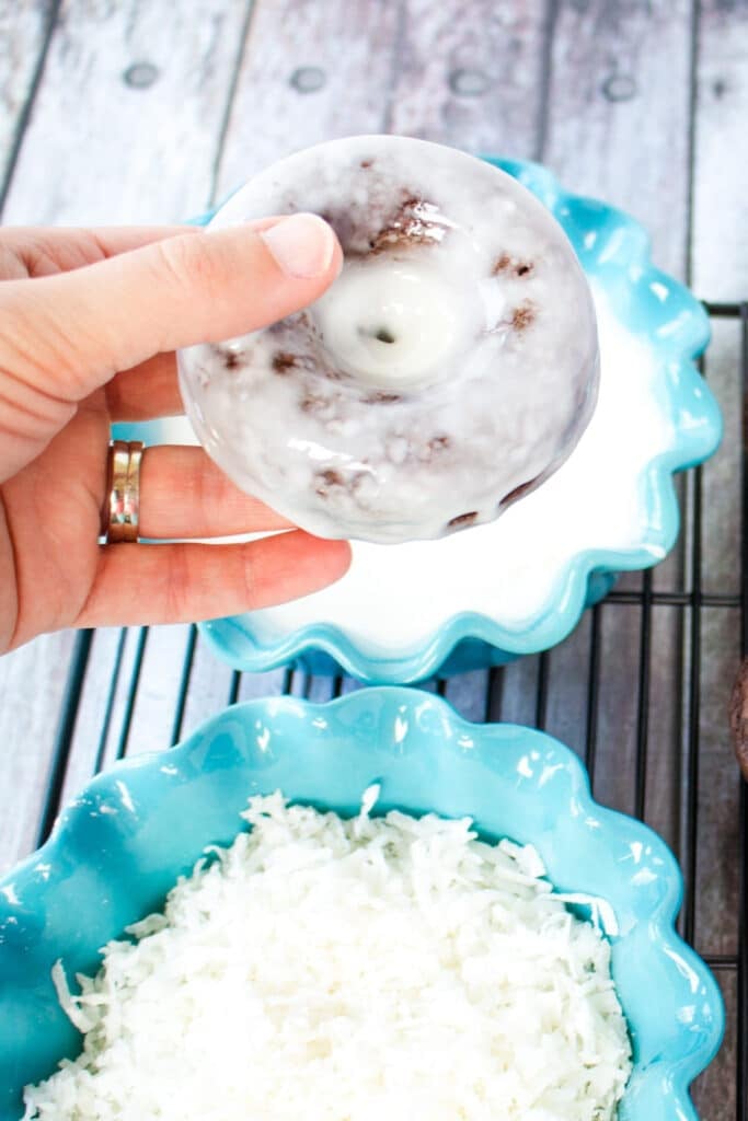 A chocolate cake donut being dipped in glaze, with bowl of coconut on the side.