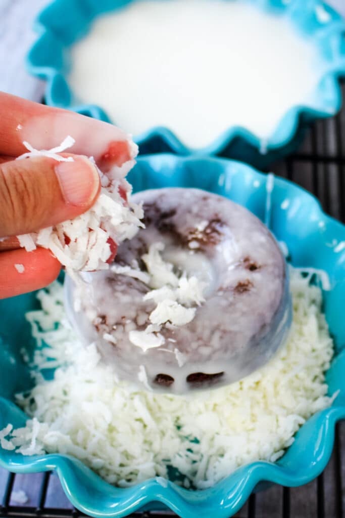 Glazed chocolate donut being dipped in coconut and coconut being sprinkled on the top.