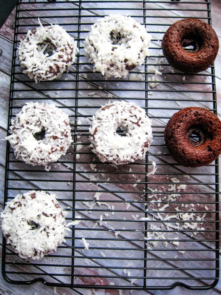 Five coconut glazed chocolate donuts on a cooling rack with two unglazed chocolate donuts on the side.