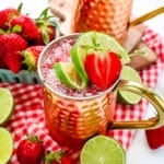 Two strawberry Moscow mules topped with fresh strawberries and lime slices with fresh strawberries and limes on the side for garnish.