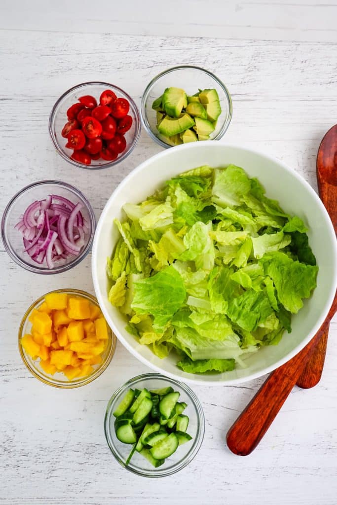 Large bowl of romaine lettuce with smaller bowls of individual ingredients to make mango chicken salad.
