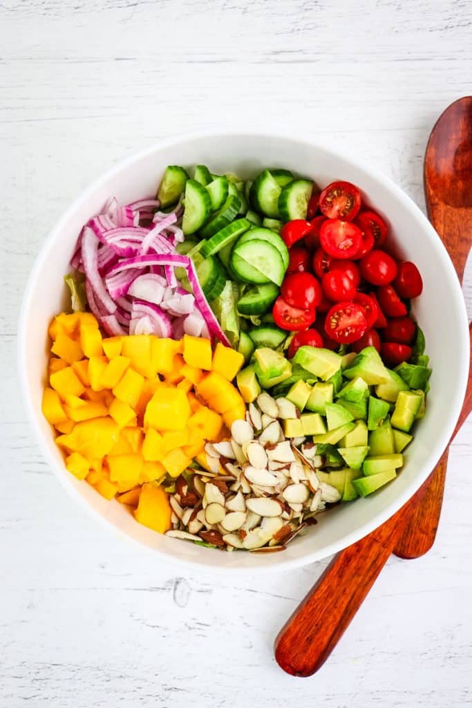 Mango chicken salad ingredients in a large white bowl with wooden utensils on the side.