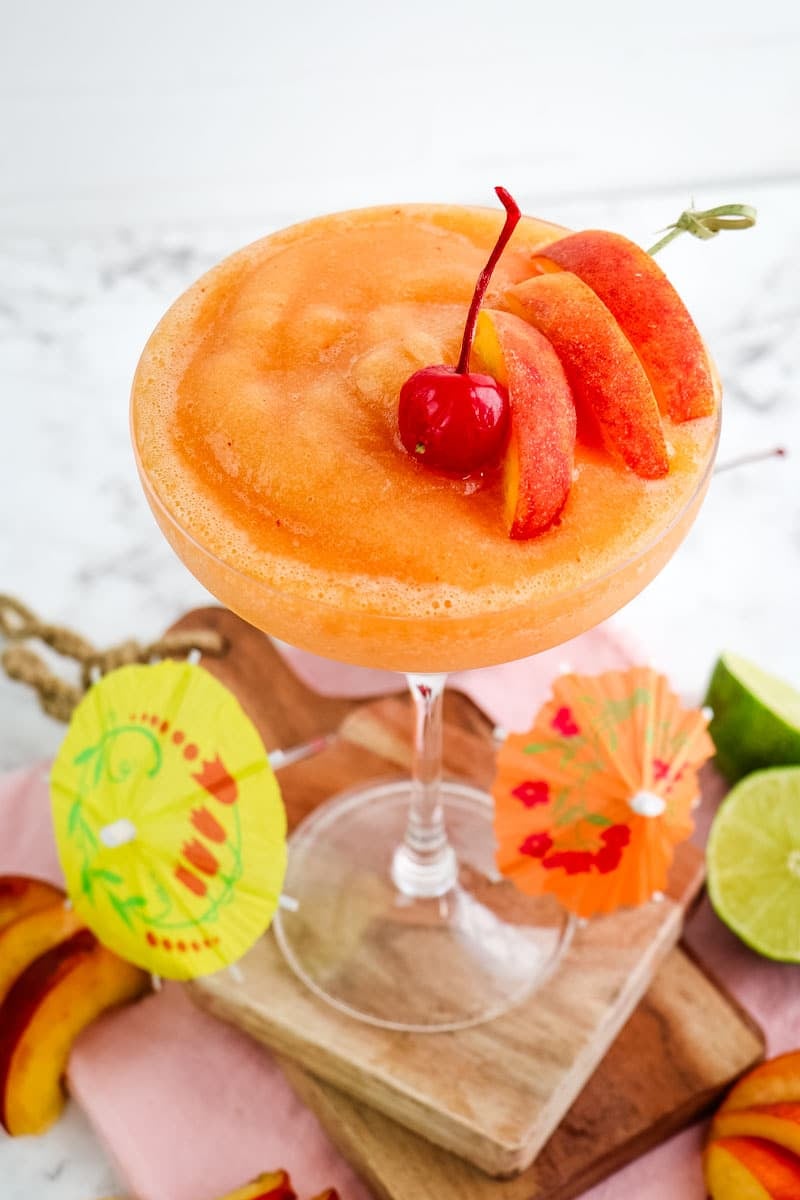 A frozen peach daiquiri topped with sliced peaches and a maraschino cherry with paper umbrellas and peach slices on the side for garnish.