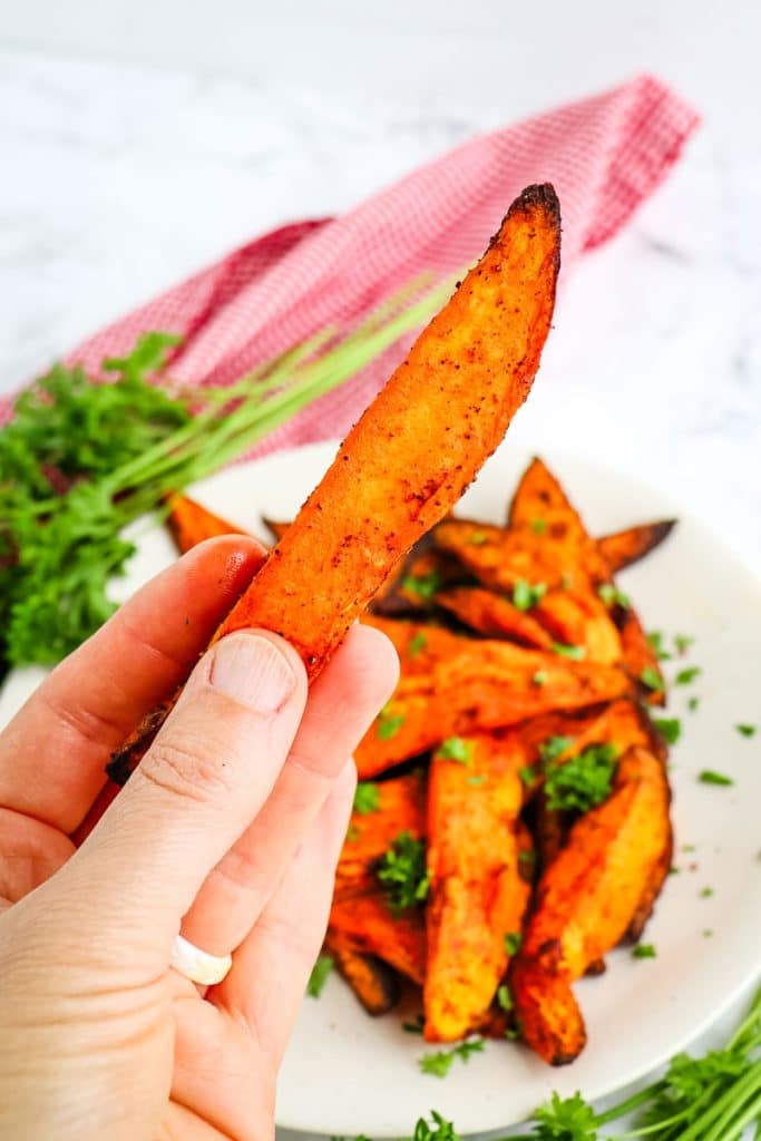 A single sweet potato wedge held in hand, with a plate of air fryer sweet potato wedges on a plate in the background.