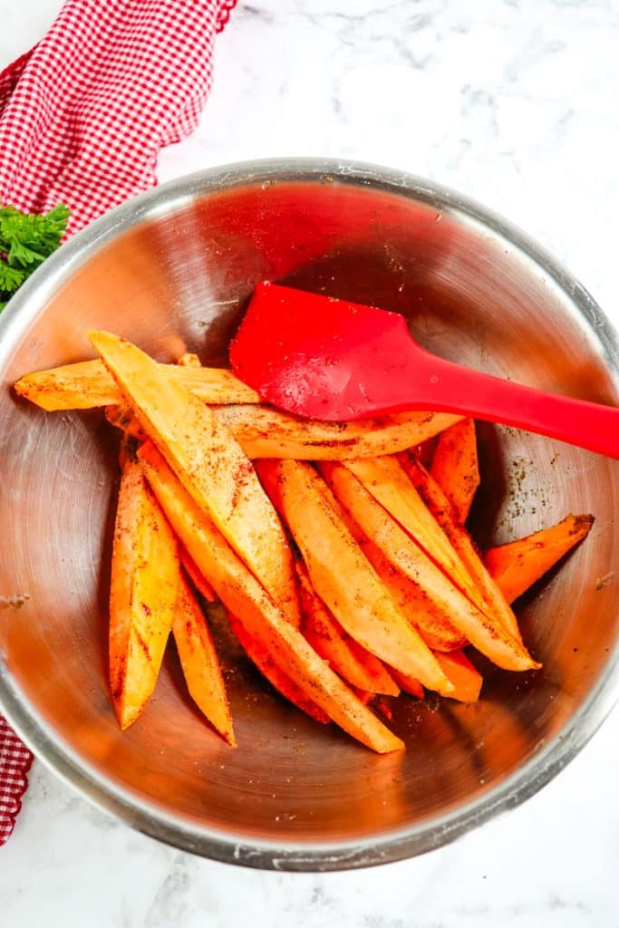 Sweet potato wedges in a mixing bowl being tossed together with spices and oil with a rubber scraper.