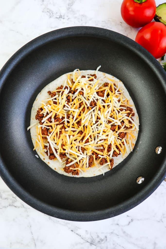 A tortilla topped with seasoned ground beef and shredded cheese in a skillet.