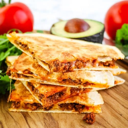 A stack of four beef quesadilla tacos with tomatoes and avocado in the background.