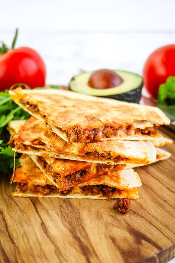 A stack of four beef quesadilla tacos with tomatoes and avocado in the background.