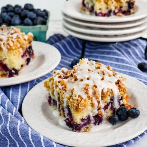 Three slices of blueberry sour cream coffee cake on white plates with fresh blueberries on the side.