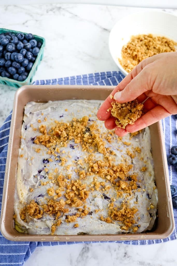 A streusel topping being sprinkled over the top of the blueberry coffee cake batter before being baked.