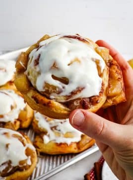 Apple pie filled cinnamon roll held in hand and topped with cream cheese frosting.