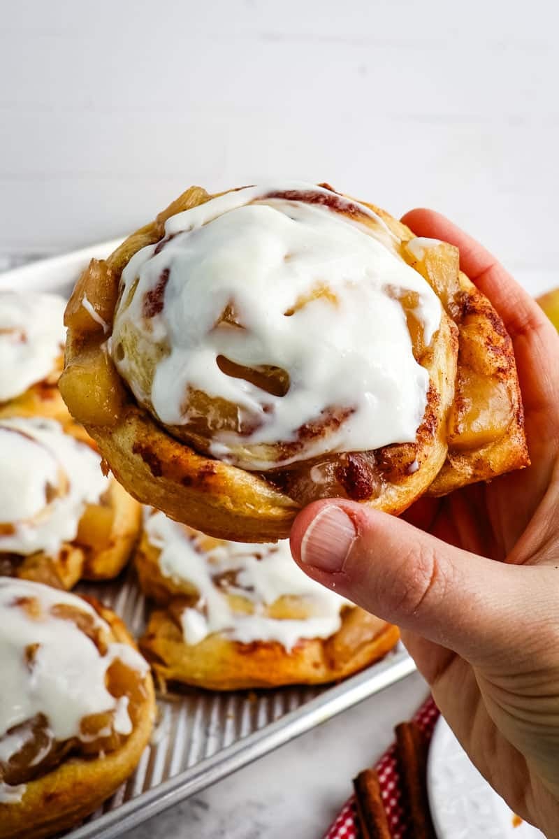 Apple pie filled cinnamon roll held in hand and topped with cream cheese frosting.