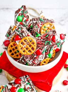 Christmas chocolate bark in a white bowl on top of a red napkin, with pieces of bark sitting on the surface below.