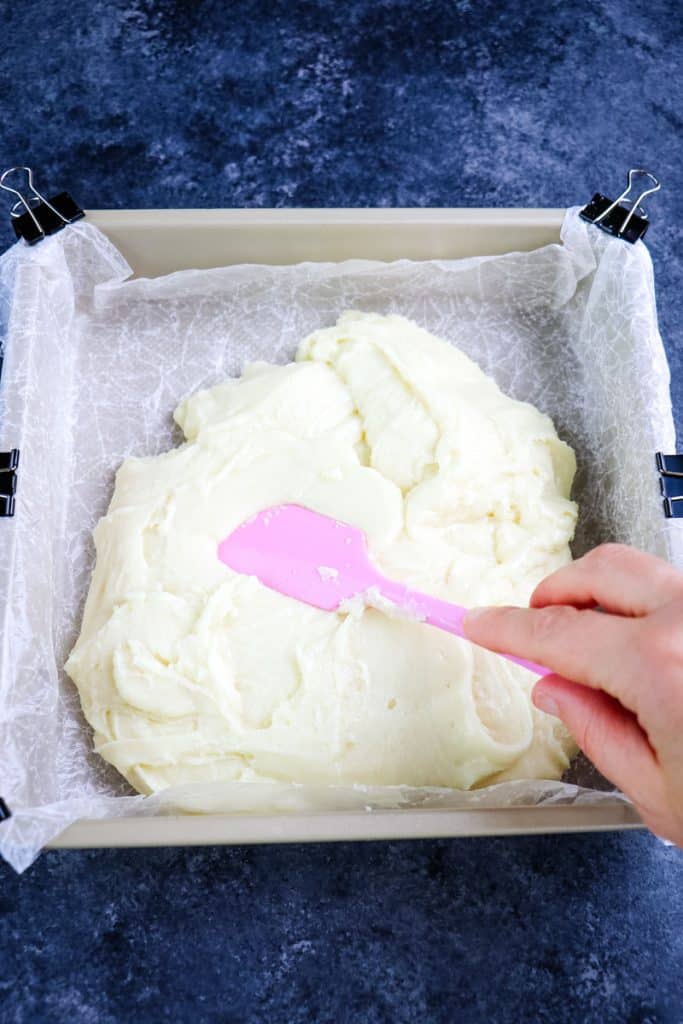 White chocolate fudge being spread into a square pan that has been lined with waxed paper.