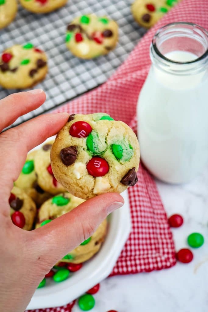 M&M cookie held in hand with milk and cookies in the background.