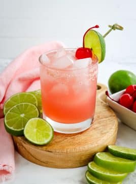 Pink margarita topped with a cherry and lime garnish with sliced limes on the side.