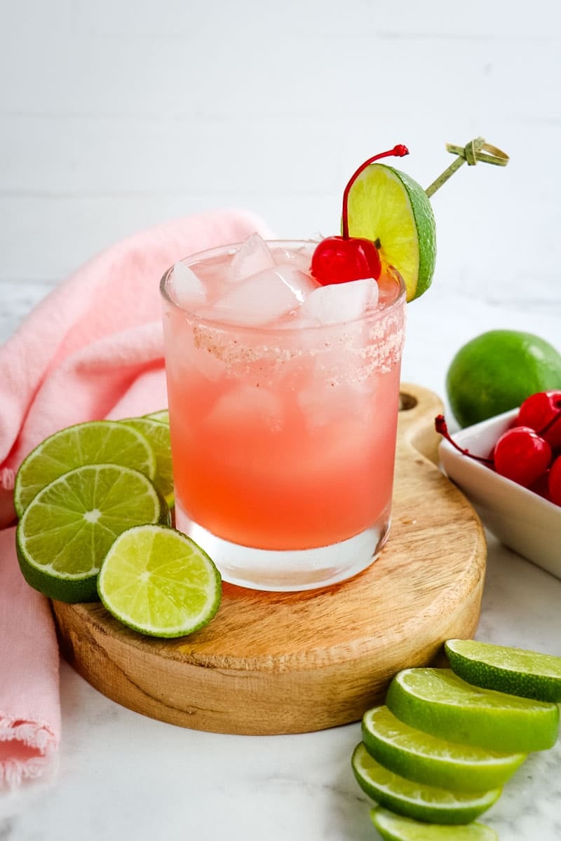 Pink margarita topped with a cherry and lime garnish with sliced limes on the side.